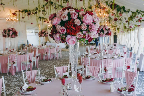 Beautiful decoration of the wedding banquet under the awning in pink, burgundy and white tones. Hall with dining tables and chairs decorated with thin cloth, bouquets, flower garlands and confetti — Stock Photo, Image