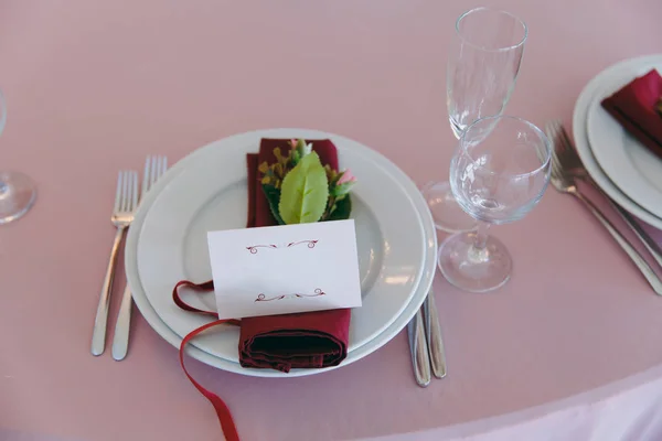 Красивое оформление свадебного банкета под навесом в розовых, бордовых и белых тонах. Накрытие стола в зале с тарелками, столовыми приборами, стаканами, салфетками и открытками, украшенными цветочными композициями — стоковое фото