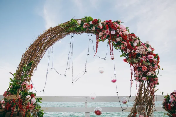 Schöne Dekoration der Hochzeitszeremonie in rosa, bordeauxroten und weißen Tönen auf einem Holzsteg. Rundbogen aus dünnen Zweigen, geschmückt mit Blumenkompositionen aus Rosen und Pfingstrosen neben dem blauen Meer — Stockfoto
