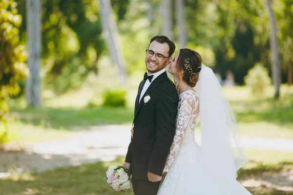Schöne Hochzeitsfotosession. der Bräutigam im schwarzen Anzug und seine Braut im weißen Spitzenkleid mit langem Federkleid, Schleier und Blumenstrauß lächeln und umarmen sich in einem großen grünen Garten an einem sonnigen Tag — Stockfoto