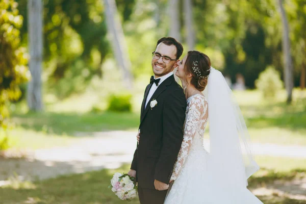 Mooie bruiloft fotosessie. De bruidegom in een zwart pak en zijn bruid in een jurk van wit kant met een lange pluim, sluier en boeket glimlach en embrace in een grote groene tuin op weathery zonnige dag — Stockfoto