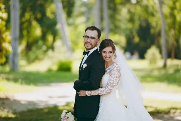 Bela sessão de casamento. O noivo em um terno preto e sua noiva em um vestido de renda branca com uma longa pluma, véu e buquê sorrir e abraçar em um grande jardim verde no dia ensolarado intempérie — Fotografia de Stock