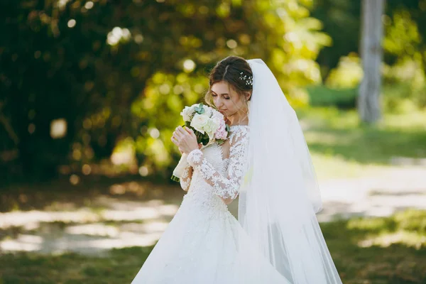 Beautiful wedding photosession. The bride in a white lace dress with a long plume, hairpin in hair, veil and bouquet of white and pink flowers in a large green garden on weathery sunny day — Stock Photo, Image