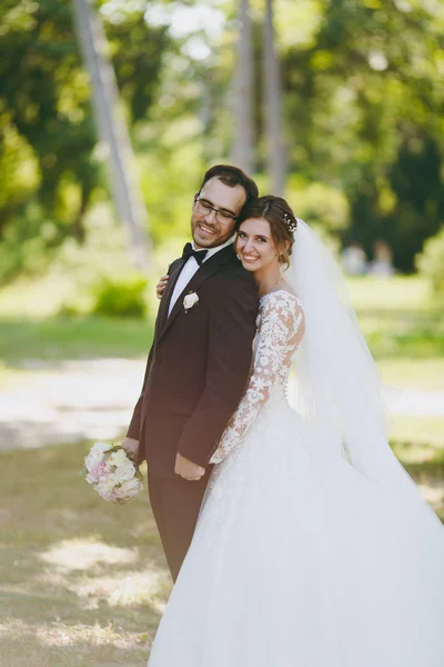 Bela sessão de casamento. O noivo em um terno preto e sua noiva em um vestido de renda branca com uma longa pluma, véu e buquê sorrir e abraçar em um grande jardim verde no dia ensolarado intempérie — Fotografia de Stock