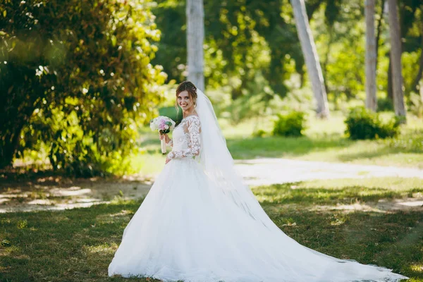 Beautiful wedding photosession. The bride in a white lace dress with a long plume, hairpin in hair, veil and bouquet of white and pink flowers in a large green garden on weathery sunny day — Stock Photo, Image