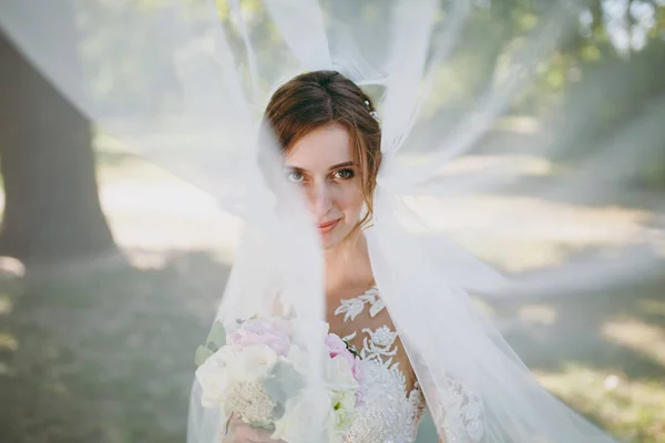 Hermosa sesión de fotos de boda. Retrato de la joven novia sonriente en un vestido de encaje blanco y ramo de flores blancas y rosadas en un velo ondulante en un gran jardín verde en el día soleado y lluvioso — Foto de Stock