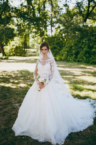 Hermosa sesión de fotos de boda. La novia en un vestido de encaje blanco con una pluma larga, horquilla en el pelo, velo y ramo de flores blancas y rosadas en un gran jardín verde en el día soleado y lluvioso — Foto de Stock