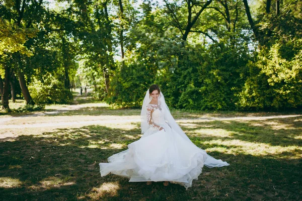Beautiful wedding photosession. The smiling bride in a white lace dress with a plume, long sleeves, hairpin in hair, white veil turns around in a large green garden on weathery sunny day — Stock Photo, Image