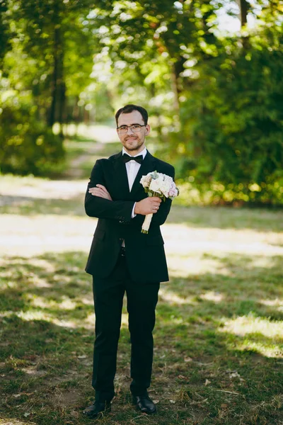 Belle séance photo de mariage. Le marié en costume noir, une chemise blanche, un nœud papillon et des lunettes avec un bouquet de fleurs blanches et roses pour la mariée dans un grand jardin vert par temps ensoleillé — Photo