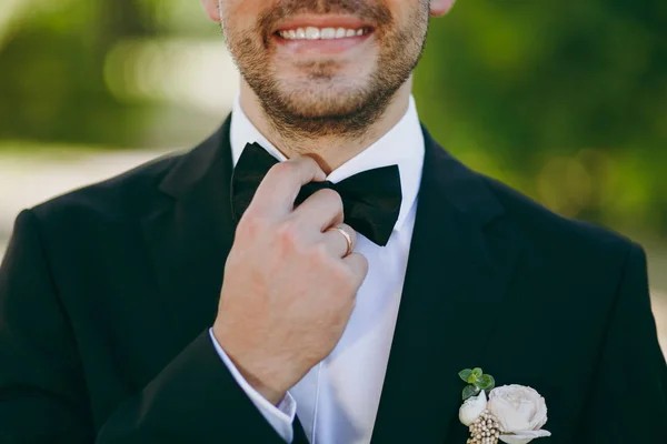 Beautiful wedding photosession. Close up The smiling groom in black suit, white shirt with boutonniere and ring on the finger straightens bow tie on a blurry green garden background — Stock Photo, Image