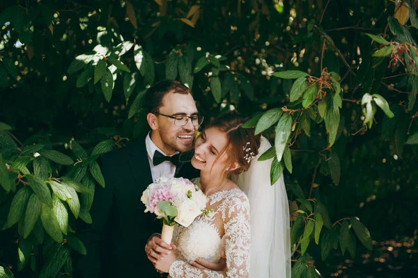 Bela sessão de casamento. Groom em terno preto, camisa branca, gravata borboleta e óculos e noiva em elegante vestido de renda com véu e buquê de flores brancas e rosa em meio aos ramos de arbusto verde — Fotografia de Stock