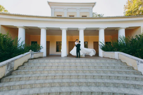 Hermosa sesión de fotos de boda. El novio en un traje negro y gafas está dando vueltas en los brazos a su joven novia linda en un vestido de encaje blanco con una pluma larga en la parte superior de las escaleras de una casa grande —  Fotos de Stock