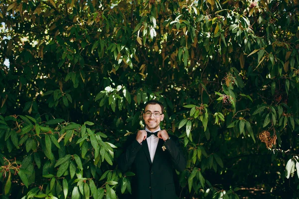 Прекрасная свадебная фотосессия. Молодой красивый улыбающийся небритый жених в черном костюме, белой рубашке и очках с бутоньеркой выпрямляет галстук-бабочку посреди ветвей зеленого куста — стоковое фото