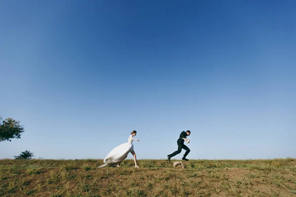 Mooie bruiloft fotosessie. Jonge charmante bruid in witte kanten jurk met lang plume loopt voor haar fianc in een zwart pak op wandeling rond het grote groene gebied tegen blauwe hemelachtergrond — Stockfoto