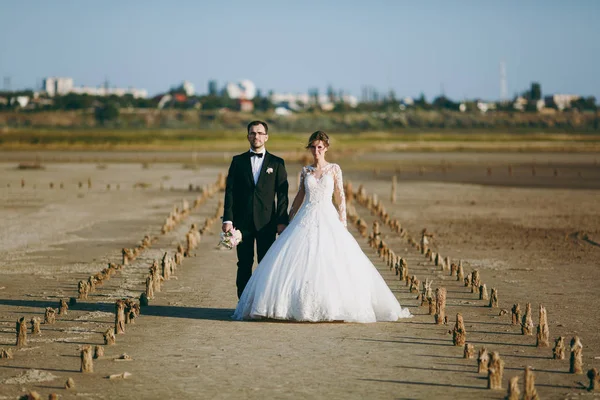 아름 다운 결혼식 photosession입니다. 꽃다발과 젊은 귀여운 신부는 해안선을 따라 산책에 절묘 한 헤어스타일으로 흰색 레이스 패턴 드레스에서와 검은 바지에 잘생긴 깎된 신랑 — 스톡 사진