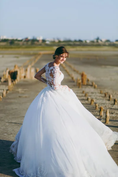 Hermosa sesión de fotos de boda. Joven linda novia elegante y bien formada en vestido de encaje blanco con una larga pluma ondeando en el viento en un paseo por la costa contra un hermoso paisaje natural — Foto de Stock