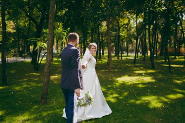 Güzel düğün photosession. Buket ve beyaz elbise onun zarif gelin resmi Mavi takım elbise ve peçe güzel hairdress güneşli bir büyük yeşil parkta bir yürüyüş ile yakışıklı damat