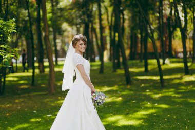 Güzel düğün photosession. Beyaz elbise ve peçe ile buket çiçek ve kurdeleler düğün güneşli büyük yeşil parkta yürüyüş ağaçta yakınındaki güzel hairdress ile zarif genç gelin