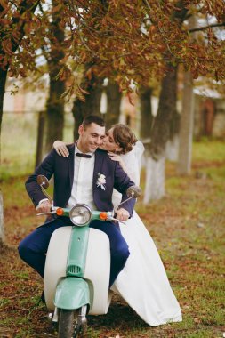 Güzel düğün photosession. Mavi resmi takım elbise ve kravat yaka çiçeği ile yakışıklı damat ile onun zarif gelin beyaz elbise ve peçe sonbahar ağaçlar yakınındaki yeşil bir vintage scooter üzerinde durur