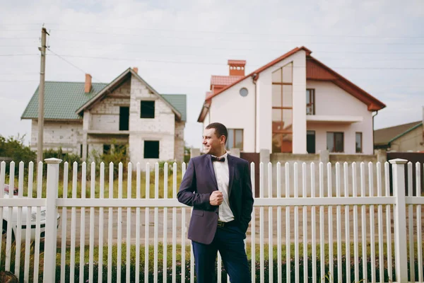 Morning preparation of the newlyweds for the wedding ceremony. The stylish smiling groom in blue formal suit, white shirt and bow tie standing on the background of fence and houses. Wedding wear
