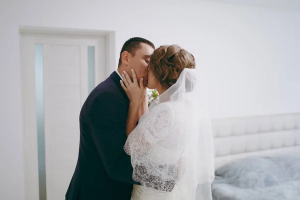 Ranní Příprava novomanželé na svatební obřad. Nevěsta a ženich snoubenec v formální oblek líbá jeho snoubenka v bílé šaty a závoj s krásný účes v pokoji — Stock fotografie
