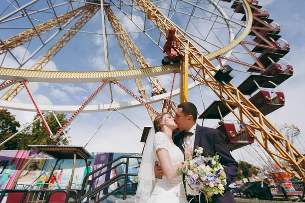 Bela sessão de casamento. Bonito noivo em um terno formal azul beija sua noiva em um vestido branco e véu com um belo hairdress contra o fundo de uma roda gigante no parque de diversões — Fotografia de Stock