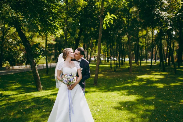 Mooie bruiloft fotosessie. Knappe bruidegom in blauwe formele pak en zijn elegante bruid in jurk en sluier met boeket met prachtige haarsnit omhelzing op een wandeling in het grote groene park op zonnige dag — Stockfoto