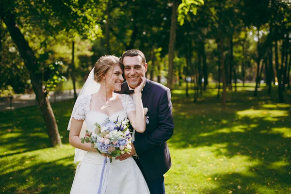 Vackert bröllop fotosession. Stilig brudgummen i blå formell kostym och sin eleganta brud i klänning och slöja med bukett med vackra frisörsal omfamning på en promenad i den stora gröna parken solig dag — Stockfoto