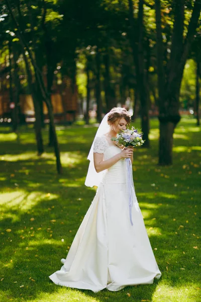 Beautiful wedding photosession. Elegant young bride in white dress and veil with beautiful hairdress with bouquet of flowers and ribbons near trees on wedding walk in the big green park on sunny day — Stock Photo, Image