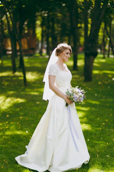 Schöne Hochzeitsfotosession. elegante junge Braut in weißem Kleid und Schleier mit schönem Haarkleid mit Blumenstrauß und Bändern in der Nähe von Bäumen beim Hochzeitsspaziergang im großen grünen Park an sonnigen Tagen — Stockfoto