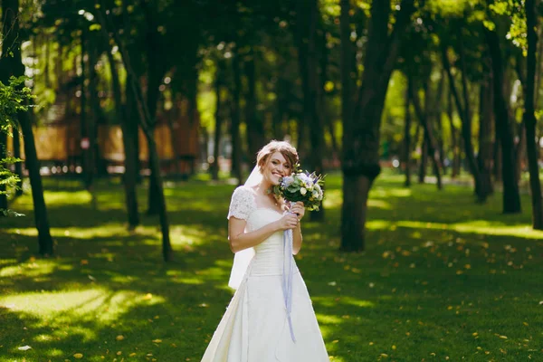 Hermosa sesión de fotos de boda. Elegante joven novia en vestido blanco y velo con hermoso peinado con ramo de flores y cintas cerca de los árboles en el paseo de bodas en el gran parque verde en el día soleado —  Fotos de Stock