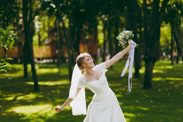 Beautiful wedding photosession. Elegant young bride in white dress and veil with beautiful hairdress with bouquet of flowers and ribbons near trees on wedding walk in the big green park on sunny day — Stock Photo, Image