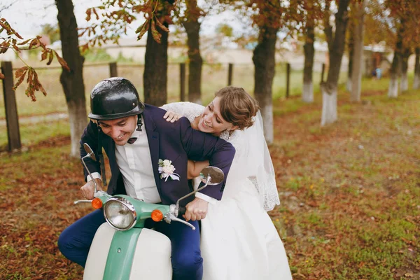 Bela sessão de casamento. Bonito noivo em azul terno formal e capacete com boutonniere e sua noiva elegante em vestido branco e véu divertindo-se em uma scooter verde perto de árvores de outono — Fotografia de Stock