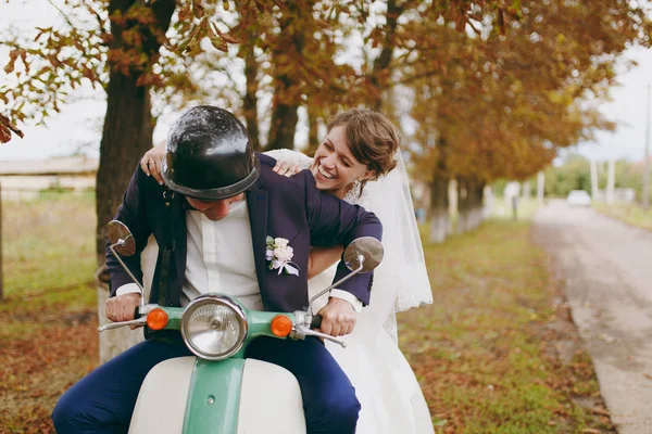 Güzel düğün photosession. Mavi resmi elbise ve kask yaka çiçeği ve onun zarif gelin beyaz elbise ve kendilerini sonbahar ağaçlar yakınındaki yeşil bir scooter üzerinde eğlenceli peçe ile yakışıklı damat — Stok fotoğraf