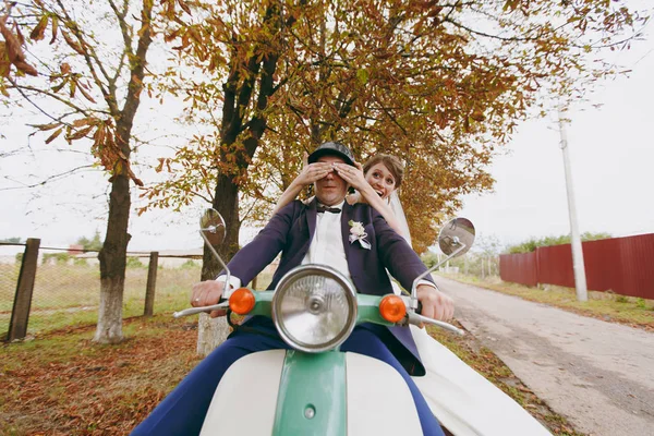Hermosa sesión de fotos de boda. Guapo novio en traje formal azul y casco con boutonniere y su novia elegante en vestido blanco y velo divertirse en una scooter verde cerca de los árboles de otoño — Foto de Stock