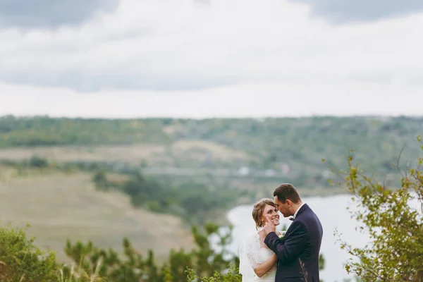 Photosession όμορφο γάμο. Όμορφος groom σε μπλε επίσημο κοστούμι και παπιγιόν με μπουτονιέρα και κομψή νύφη σε λευκό φόρεμα και πέπλο με μια όμορφη κομμωτι σε έναν περίπατο στο χωράφι κοντά στον ποταμό — Φωτογραφία Αρχείου