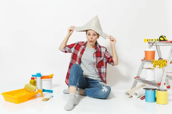 Διασκέδαση ευτυχισμένη γυναίκα με καπέλο εφημερίδα κάθεται στο πάτωμα με τα όργανα για δωμάτιο διαμέρισμα ανακαίνιση που απομονώνονται σε λευκό φόντο. Ταπετσαρία, αξεσουάρ για την κόλληση, εργαλεία ζωγραφικής. Επισκευή concept home. — Φωτογραφία Αρχείου