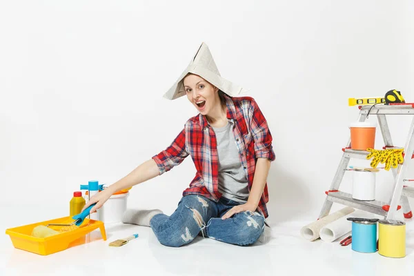 Aranyos nő ül a padlót, FESTŐHENGER, eszközök felújítás apartman elszigetelt fehér background újság kalapban. Tapéták ragasztására, festőeszközök tartozékok. Javítás otthon koncepció — Stock Fotó