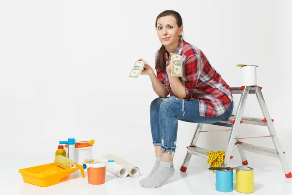 Upset female holds tight of dollars, cash few money, sits on ladder with instruments for renovation apartment isolated on white background. Wallpaper, gluing accessories painting tools. Repair concept — Stock Photo, Image