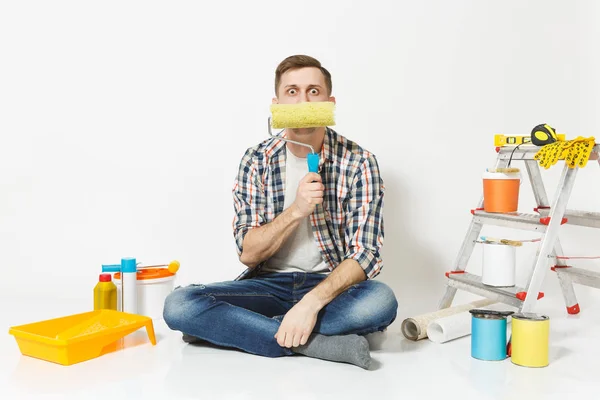 Hombre divertido sentado en el suelo con rodillo de pintura como bigote, instrumentos para la renovación habitación del apartamento aislado sobre fondo blanco. Fondo de pantalla, pegar accesorios, herramientas de pintura. Reparación concepto hogar . — Foto de Stock