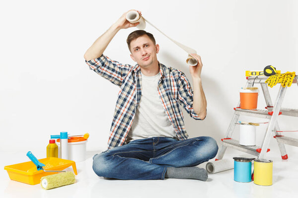 Confused perplexed man sitting on floor with roll of wallpaper above head, instruments for renovation apartment isolated on white background. Gluing accessories, painting tools. Repair home concept.