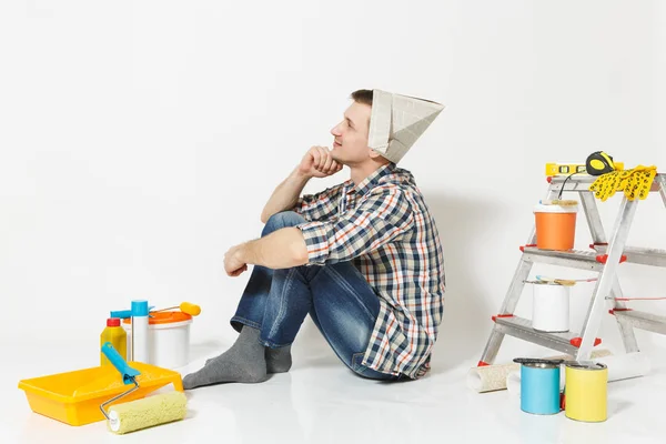 Joven en sombrero de periódico sentado en el suelo con instrumentos para la renovación habitación de apartamentos aislado sobre fondo blanco. Fondo de pantalla, pegar accesorios, herramientas de pintura. Reparar concepto de casa. Vista lateral . — Foto de Stock