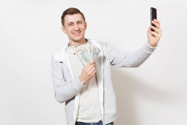 Genç gülümseyen yakışıklı t-shirt, boynuna kulaklıklarla ışık kazak bekle paket dolar, nakit para ve resimlerin kendini beyaz arka plan üzerinde izole telefon. Başarı kavramı