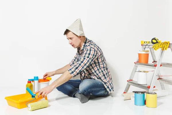 Συγχέεται σοκαρισμένος άνθρωπος στην εφημερίδα καπέλο εξάπλωση τα χέρια, κάθεται στο πάτωμα με τα όργανα για διαμέρισμα ανακαίνιση που απομονώνονται σε λευκό φόντο. Ταπετσαρία κόλλημα αξεσουάρ εργαλεία. Επισκευή concept home. — Φωτογραφία Αρχείου