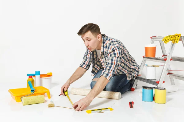 Νεαρός άνδρας κάθεται στο πάτωμα με ρολό ταπετσαρία, μολύβι, μέτρο ταινία, όργανα για ανακαίνιση Διαμέρισμα Δωμάτιο απομονωθεί σε λευκό φόντο. Κόλλημα αξεσουάρ, εργαλεία βαφής. Επισκευή concept home — Φωτογραφία Αρχείου