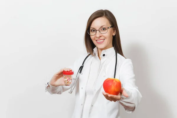 Sonriente joven doctora confiada con estetoscopio, gafas aisladas sobre fondo blanco. La doctora en bata médica sostiene la botella con pastillas, manzana roja. Personal sanitario, concepto de medicina . — Foto de Stock