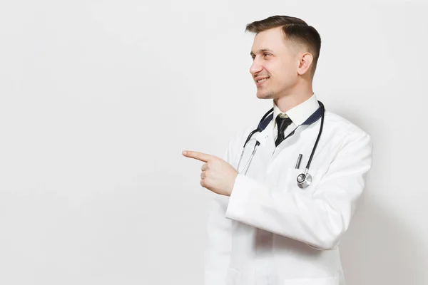 Sorridente bel giovane medico isolato su sfondo bianco. Medico maschio in abito medico, stetoscopio indicando dito indice da parte su spazio copia. Personale sanitario, salute, concetto di medicina . — Foto Stock