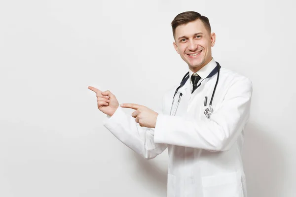 Sorridente bel giovane medico isolato su sfondo bianco. Medico maschio in abito medico, stetoscopio indicando dita indice da parte su spazio copia. Personale sanitario, salute, concetto di medicina . — Foto Stock