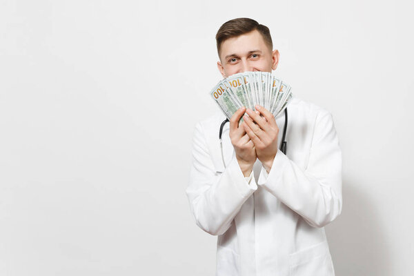 Smiling young doctor man isolated on white background. Male doctor in medical uniform, stethoscope holding bundle of dollars, banknotes, cash money. Healthcare personnel, health, medicine concept.