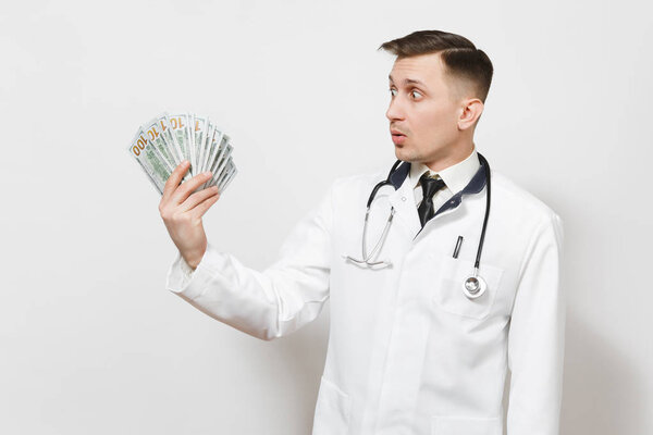 Shocked young doctor man isolated on white background. Male doctor in medical uniform, stethoscope looking on bundle of dollars, banknotes, cash money. Healthcare personnel, health, medicine concept.